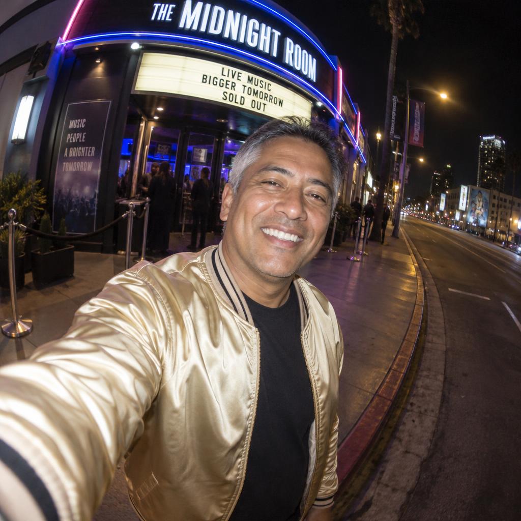 Fisheye Night-Out Snapshot: A close selfie at the edge of a neon-lit concert venue entrance, his broad grin near the center and his outstretched arm entering the bottom…