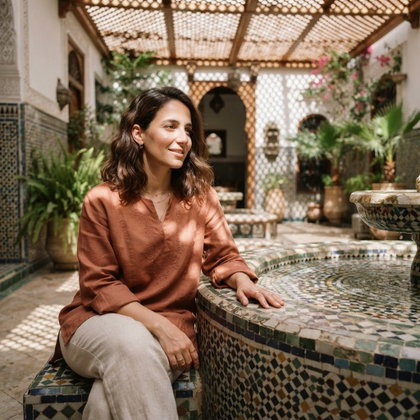 A single subject sits beside a mosaic fountain in a quiet tiled courtyard, one hand resting on its stone edge. A loose terracotta tunic and simple linen trousers echo the warm architecture. Sharp midday sunlight casts patterned shadows from an overhead lattice; off-center waist-up composition, refined travel-editorial photography.