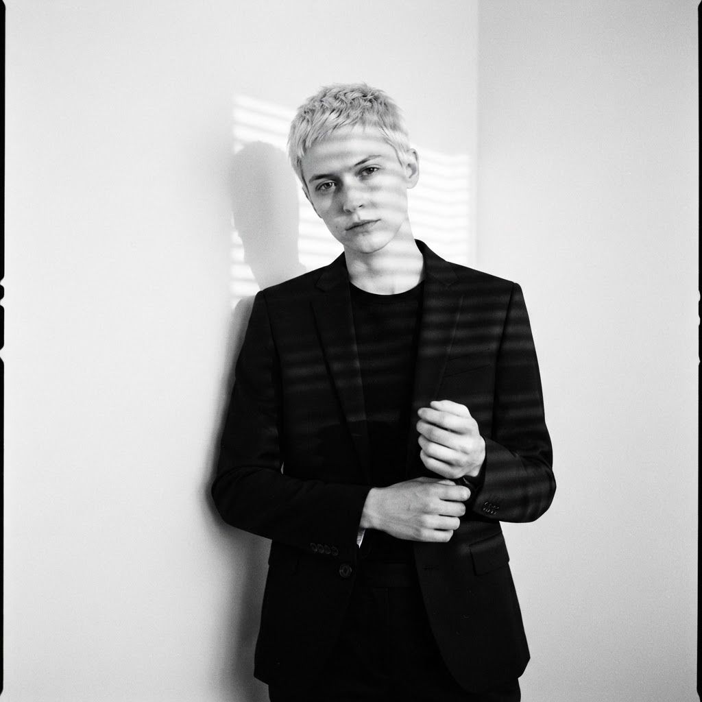 Venetian Blind Shadows: Half-body portrait in a minimalist noir apartment corner, one visible person in a fitted black suit standing close to a blank wall.